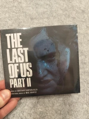 The Last of Us Part II soundtrack - Soundtrack till spelet The Last of Us Part II med musik av Gustavo Santaolalla och Mac Quayle. Omslaget är svart med vit text och har en bild på en gitarr. Perfekt för dig som gillar spelmusik och samlarprylar från PlayStation-universumet.