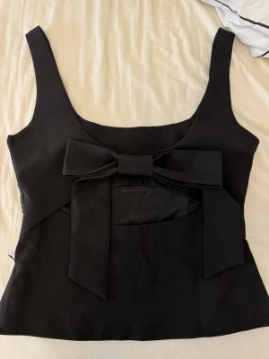 Svart topp med stor rosett - Snygg svart linnetopp med breda axelband och en stor dekorativ rosett framtill. Toppen har en figurnära passform och är tillverkad i ett slätt tyg som ger en stilren look. Perfekt att styla med jeans eller kjol för en trendig outfit.