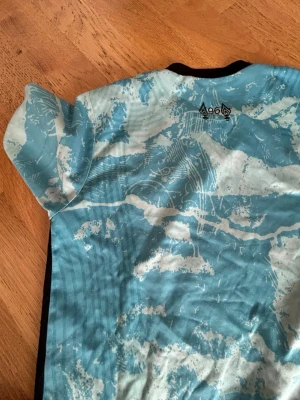 Blå sporttröja från AGOA - Säljer en blå sporttröja från AGOA med korta ärmar och ett unikt mönster i vitt och ljusblått. Tröjan har svarta detaljer längs sidorna och är tillverkad i ett lätt och funktionellt material som passar perfekt för träning.