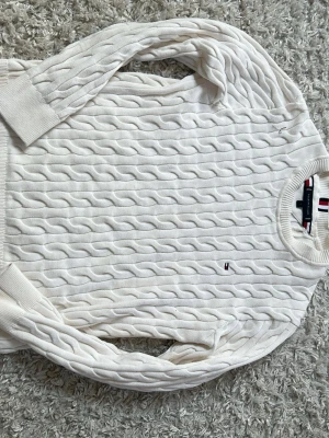 Vit kabelstickad tröja Tommy Hilfiger - Säljer en vit kabelstickad tröja från Tommy Hilfiger. Tröjan har rund halsringning, ribbade muddar och klassisk kabelstickad struktur. Perfekt för dig som gillar stilrena och tidlösa plagg. Diskret logodetalj på bröstet.