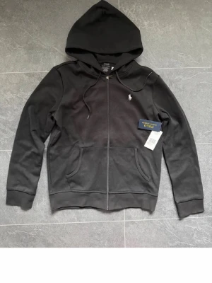 Svart hoodie från Polo Ralph Lauren - Snygg svart hoodie med dragkedja från Polo Ralph Lauren. Klassisk design med vit broderad logga på bröstet, huva med dragsko och två stora fickor framtill. Tillverkad i mjukt bomullsmaterial som känns skönt mot huden.