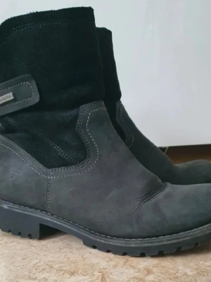 Svarta fodrade boots från Superfit - Säljer ett par svarta boots från Superfit med grov sula och mjukt, varmt foder. Ovandelen är i mocka och nubuck med dekorativa sömmar och kardborreband på sidan. Perfekta för kalla dagar när du vill ha både stil och komfort. Storlek 38