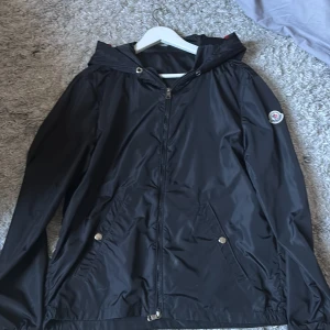 Moncler windbreaker - Säljer en svart vindjacka från Moncler med huva och dragkedja framtill. Jackan har två fickor med tryckknappar och Moncler-logga på vänster ärm. Materialet är glansigt och lätt, perfekt för att skydda mot vind. Röd och vit detalj på huvan baktill.