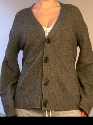 Grå oversized kofta med stora knappar, från Nelly - Mysig grå kofta i oversized modell med fyra stora svarta knappar framtill. Koftan har djup V-ringning, långa ärmar och ribbade muddar. Perfekt att slänga över en topp för en chill look.