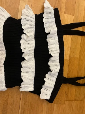 Svart ribbad topp med vita volanger - Trendig svart ribbad topp med breda axelband och dekorativa vita virkade volanger framtill. Materialet känns stretchigt och bekvämt, perfekt för att skapa en cool och unik look. Volangerna ger toppen en söt och lekfull touch.