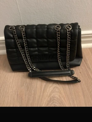 Steve Madden väska - Snygg svart axelväska i quiltad design med grova silverfärgade kedjor som axelrem. Väskan har två stora fack och är gjord i ett mjukt, läderliknande material. Perfekt för dig som gillar trendiga accessoarer med lite edge.