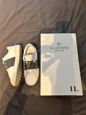 Valentino Garavani vita sneakers - Snygga vita sneakers från Valentino Garavani med bred grå remsa över sidan och coola nitar på hälen. Skorna är i läder och har klassisk låg profil med vita snören och gummisula. Perfekta för dig som gillar stilrena och exklusiva detaljer. Skorna är storlek 39 men passar mycket bättre på en 40.