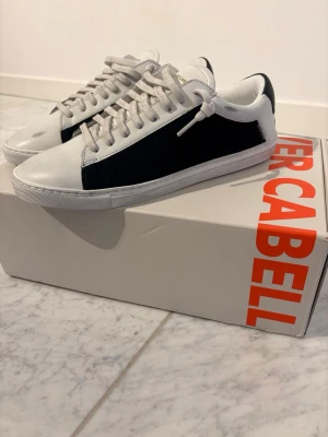Vita och mörkblå sneakers Oliver Cabell - Snygga sneakers från Oliver Cabell i vitt och mörkblått. materialet är nubuck och mocka. Skorna är helt nya. Köpta från Oliver Cabells hemsida i USA nu i april. Tyvärr för små för sonen. Orderbekräftelse/kvitto kan visas för den som önskar. Klassisk låg modell med vita snören och diskret guldlogga på plösen. Skorna har en stilren design med kontrasterande sidopaneler och vit sula. nypris ligger på 2000 kr exkl frakt och tull som gick på 600 kr.  Hör av er vid frågor eller om fler bilder önskas. 