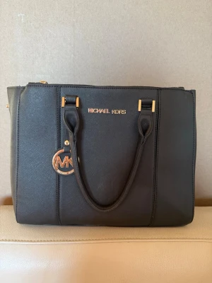 Svart handväska från Michael Kors - Stilren svart handväska från Michael Kors med guldfärgade detaljer och logotyp. Väskan har två handtag, dragkedja upptill och ett hängande MK-emblem i metall. Tillverkad i tåligt konstläder med en klassisk, fyrkantig form.