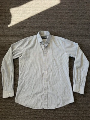 Skjorta - Klassisk vit skjorta med tunna blå ränder. Skjortan har lång ärm, bröstficka och knappar framtill. Tillverkad i mjuk bomull och passar perfekt till både jeans och chinos.