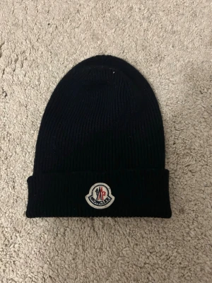 Svart Moncler mössa med logga - Hej, Säljer en svart ribbstickad mössa från Moncler med uppvikt kant och klassisk Moncler-logga framtill. Mössan är tillverkad i mjukt material och har en stilren, enkel design som passar perfekt till höst och vinter. 