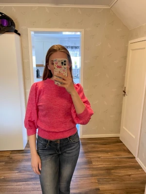Rosa stickad tröja med puffärm - Säljer en stickad tröja i starkt rosa med puffärmar och ribbade muddar. Tröjan har en något kortare passform och rund halsringning. Ärmen går även att dra upp lite för att få den kortare. Perfekt för dig som vill ha en färgklick i garderoben och gillar trendiga detaljer.
