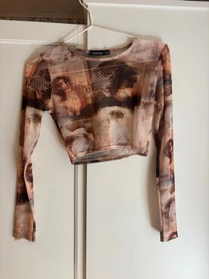 Mönstrad croppad topp från Boohoo - Trendig croppad topp från Boohoo med lång ärm och abstrakt mönster i bruna, beige och svarta toner. Tunn mesh-liknande polyester som ger en cool och edgy vibe. Tråden i en söm längst ner är delvis borta (tredje bilden) men annars är den i bra skick. 