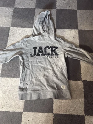 Grå hoodie från Jack & Jones - Snygg grå hoodie från Jack & Jones med stor svart logga på bröstet. Klassisk modell med huva och långa ärmar. Tillverkad i mjukt bomullsmaterial som känns skönt mot huden. Perfekt för en avslappnad och trendig stil.