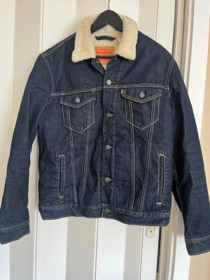 Levi's mörkblå jeansjacka med foder - Snygg mörkblå jeansjacka från Levi's med vitt teddyfoder i kragen. Klassisk modell med två bröstfickor med lock och knappar, samt två sidofickor. Kontrastsömmar och knappstängning framtill ger en tidlös look.