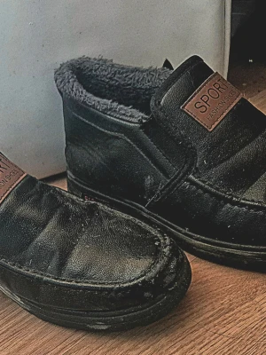Svarta fodrade skor SPORT - Mysiga svarta skor från SPORT med mjukt grått foder och skön passform. Utsidan är i läder och insidan är fodrad med fluffigt material för extra värme. Perfekta för kalla dagar  Klassisk design med en liten etikett framtill.