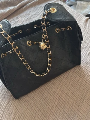 Svart fake chanel  handväska - Säljer en klassisk svart handväska, kopia av den trendiga Chanel väskan. Med guldfärgade kedjedetaljer. Väskan har en rymlig form och eleganta detaljer som gör den till ett statement piece. Perfekt för dig som vill ha något tidlöst och lyxigt. Aldrig använt. 