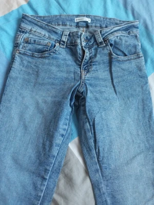 Ljusblå jeans från Gina Tricot  - Säljer ett par klassiska bootcut, ljusblå jeans från Gina Tricot i storlek 40, S/M. De har fyra fickor, låg midja och utsvängda ben. Jeansen är tillverkade i ett mjukt denimt bayg och har snygga detaljer med nitar vid fickorna. Pe barfekta till en avslappnad look.