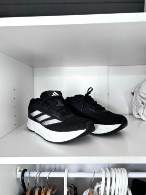 Adidas Skor  - Snygga svarta sneakers från Adidas med klassiska vita ränder på sidan och vit sula. Ovandelen är i mesh för bra andningsförmåga och skon har en sportig, modern look. Perfekta för dig som gillar streetstyle och vill ha bekväma skor till vardags. Inte så smutsiga bara lite smuts som går bort med vatten. Fraktas timmen de köps. 