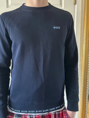Blå sweatshirt från BOSS - Stilren blå sweatshirt från BOSS med rund halsringning och diskret BOSS-logga på bröstet. Ribbstickade muddar vid ärmslut och nederkant, samt BOSS-tryck längs nederkanten. Perfekt till jeans eller joggers.
