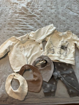 Sött babyset med bodys och dregglisar - Ett gulligt babyset med två långärmade bodys i beige toner, en med texten 'hello world' och en med Nalle Puh-tryck. Till setet hör tre ribbade dregglisar i beige, brun och grå samt ett par grå byxor med små fjärilar. Mjuka material, perfekt för små barn.