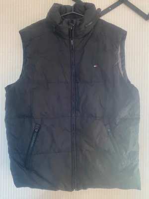 Svart dunväst från Tommy Hilfiger - Stilren svart dunväst från Tommy Hilfiger med hög krage och två dragkedjeförsedda fickor framtill. Västen har diskret logga på bröstet och quiltad design som ger en modern look. Perfekt att slänga över en hoodie eller tröja för extra värme.