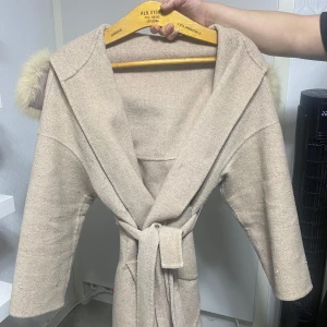 Beige kappa med pälskrage och bälte  - Snygg beige kappa med stor huva och fluffig pälskrage. Jackan har knytskärp i midjan, stora fickor och trekvartsärmar. Perfekt för dig som vill ha en stilren och mysig look under höst och vinter. Man får med dens puffar som man kan lägga på och ta av. Storlek S/M