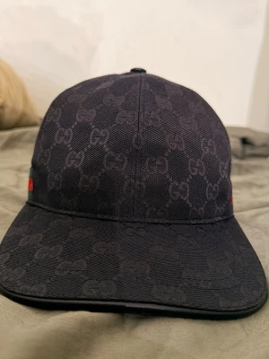 Svart Gucci keps med GG-mönster - Säljer en svart keps från Gucci med det klassiska GG-monogrammönstret över hela kepsen. På sidan finns en grön och röd rand som ger en ikonisk touch. Kepsen har böjd skärm och är tillverkad i ett slitstarkt tygmaterial.
