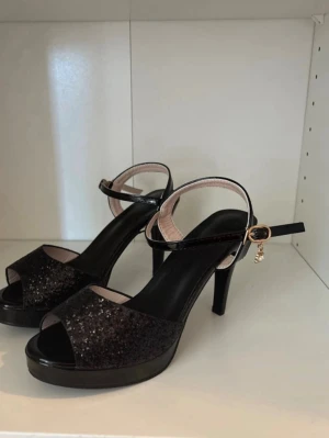 Svarta glittriga pumps med platå - Säljer ett par svarta pumps med glittrig ovandel, öppen tå och hög klack. Skorna har en smal ankelrem med guldfärgat spänne och liten hängande dekoration. Platåsulan ger extra höjd och komfort. Perfekta för dig som vill sticka ut lite extra.