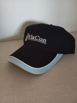 Svart keps från ViaCon med ljusblå kant - Snygg svart keps från ViaCon med broderad logga framtill. Kepsen har en böjd skärm med ljusblå kant och vit detalj längs skärmens ytterkant. Tillverkad i ett mjukt tygmaterial och har klassisk passform.
