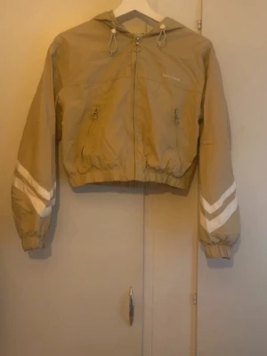 Beige vindjacka  - Snygg beige vindjacka från Extr.Street med vita detaljer på ärmarna och dragkedjor med ringar. Jackan har huva med dragsko, två fickor framtill och resår i midjan och vid ärmsluten. Perfekt för vår och höst.