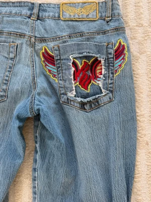 Ljusblå bootcut jeans med brodyr - Unika ljusblå jeans från Antik Denim med bootcut passform. Bakfickan har en stor broderad detalj med röda och gula vingar samt slitningar. Klassisk femficksmodell och cool vintagekänsla. Perfekta för dig som vill sticka ut med detaljer och färg.