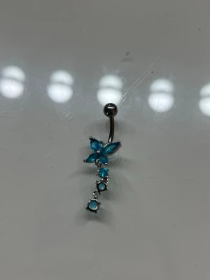 Navelpiercing med blå fjäril - Snygg navelpiercing i silverfärgad metall med hängande dekoration i form av en blå fjäril och flera blå stenar. Fjärilen har fyra vingar av blå glittrande stenar och smycket har en böjd stav. Perfekt för dig som vill ha något unikt och glittrigt.