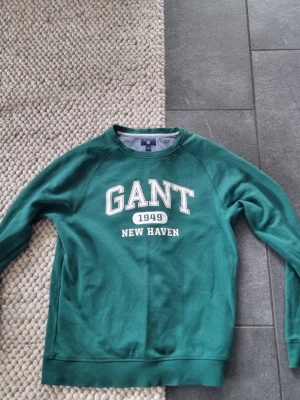 Grön sweatshirt från GANT - Mörkgrön sweatshirt från GANT med vit text 'GANT 1949 NEW HAVEN' på bröstet. Klassisk rund halsringning och långa ärmar. Perfekt för en avslappnad och sportig stil. Mjuk bomullskvalitet och ribbade muddar vid ärmslut och nederkant.