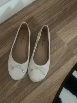 Vita ballerinaskor med rosett - Säljer ett par klassiska vita ballerinaskor från S. Oliver. Skorna har rund tå, dekorativ rosett framtill med guldfärgade detaljer och en enkel, stilren design. Perfekta för dig som gillar en clean och tidlös look.