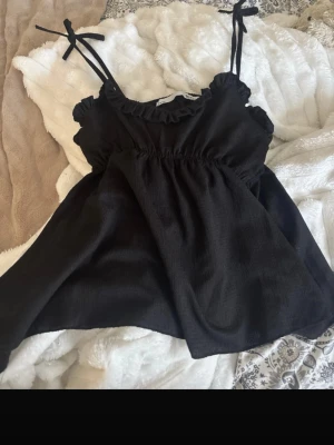 Svart linnetopp med volang och knyt - Söt svart linnetopp med volangkant vid ringningen och smala axelband som knyts upptill. Luftig modell med lätt rynk under bysten för en avslappnad look. Perfekt till jeans eller kjol för en trendig stil.