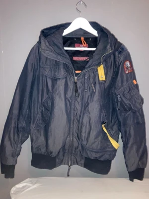 Svart vindjacka från Parajumpers - Cool svart vindjacka från Parajumpers med huva, flera fickor och ikoniska gula detaljer. Jackan har patch på ärmen och ribbade muddar. Perfekt för dig som vill ha en sportig och urban stil.