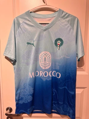 Blå fotbollströja Marocko Puma - Snygg fotbollströja från Puma i ljusblått till mörkblått ombre-mönster. Marockos landslagsemblem på bröstet och trycket 'MOROCCO' framtill. Tillverkad i lätt och ventilerande funktionsmaterial, perfekt för träning eller match.