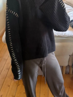 Svart stickad tröja med vita detaljer - Säljer en svart ribbstickad tröja med vita dekorativa sömmar längs ärmarna. Tröjan har en avslappnad passform och långa ärmar. Perfekt för dig som gillar en unik twist på klassisk stickat.