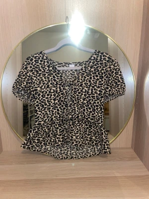 Leopardmönstrad blus med rosetter  - Säljer en leopardmönstrad blus med korta ärmar och rosetter framtill. Toppen har volang nedtill och en v-ringad urringning. Perfekt för dig som vill sticka ut lite extra.skriv för fler bilder, pris kan diskuteras!💗