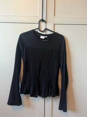 Svart långärmad topp med volang - Säljer en svart långärmad topp från Gina Tricot. Toppen har smockad front och volangkant nedtill som ger en snygg siluett. Tillverkad i mjukt bomullsmaterial och passar perfekt till jeans eller kjol.