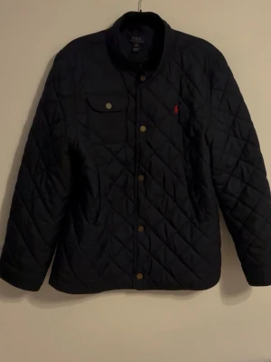 Svart quiltad pufferjacka Polo Ralph Lauren - Stilren svart quiltad pufferjacka från Polo Ralph Lauren med röd broderad logga på bröstet. Jackan har tryckknappar, en bröstficka med lock och två sidofickor. Perfekt för kalla dagar och har en klassisk, tidlös look. Storlek XL men passar M