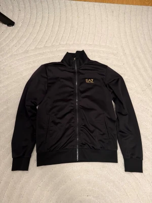 Svart ziptröja och byxa från EA7 Emporio Armani - Snygg svart ziptröja från EA7 Emporio Armani med guldfärgad logga på bröstet och guldfärgade ränder på ryggen. Tröjan har hög krage, långa ärmar och ribbade muddar. Tillverkad i ett slätt, sportigt material som känns bekvämt och stilrent.249kr för tröjan och 199 för byxan