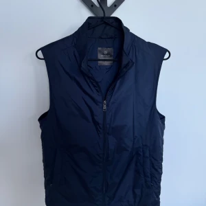 Mörkblå väst från Berkeley - Snygg mörkblå dunväst från Berkeley med hög krage och dragkedja framtill. Västen har en stilren design och är tillverkad i ett glansigt material som ger en modern look. Perfekt att slänga över en hoodie eller tröja för extra värme.