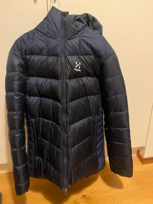 Mörkblå dunjacka från Haglöfs - Snygg mörkblå dunjacka från Haglöfs med huva och hel dragkedja framtill. Jackan har ett puffigt, quiltat mönster och Haglöfs-logga på bröstet. Perfekt för kalla vinterdagar och riktigt varm tack vare dunfyllningen.