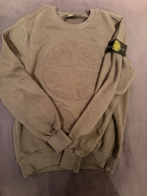 Olivgrön sweatshirt från Stone Island - Säljer en olivgrön sweatshirt från Stone Island med det klassiska broderade logotypmärket på ärmen och stort Stone Island-emblem på bröstet. Tröjan har rund hals, ribbade muddar och är tillverkad i mjuk bomull. Ny pris ~2100