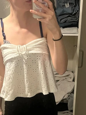 Vit broderad linnetopp - Söt vit linnetopp med broderade hålmönster och dekorativ knut framtill. Toppen har tunna axelband och en luftig, flowy passform som passar perfekt till sommaren.