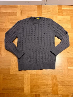 Grå kabelstickad tröja från polo Ralph Lauren - Snygg grå kabelstickad tröja från polo Ralph Lauren med rund halsringning och diskret logga broderad på bröstet. Tröjan har långa ärmar och ribbade muddar vid ärmslut och nederkant. Perfekt för dig som gillar klassisk och stilren design.