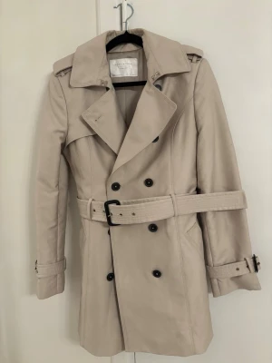 Beige trenchcoat från Zara Woman - Stilren beige trenchcoat från Zara Woman med dubbelknäppning, bred krage och bälte i midjan. Jackan har svarta knappar, axelklaffar och justerbara spännen vid ärmsluten. Perfekt för höst och vår, tillverkad i ett slätt tyg.