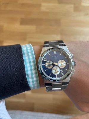 Tissot blå kronograf herrklocka - Snygg Tissot herrklocka med blå urtavla och tre silverfärgade subdials. Klockan har ett robust armband i rostfritt stål och klassisk rund form. Perfekt accessoar för dig som gillar stilrena detaljer och schweizisk design.